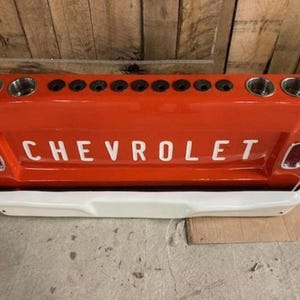 Op de afbeelding: Een rode Chevrolet-truckachterklep met het woord "CHEVROLET" in witte letters. De achterklep heeft twee bekerhouders aan elk uiteinde en een reeks kleinere gaten in het midden. De onderkant is wit.