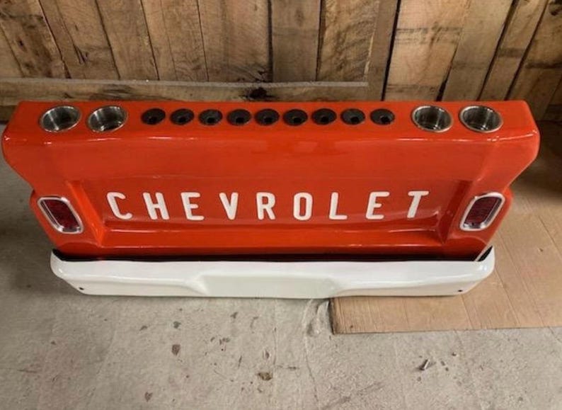 Op de afbeelding: Een rode Chevrolet-truckachterklep met het woord "CHEVROLET" in witte letters. De achterklep heeft twee bekerhouders aan elk uiteinde en een reeks kleinere gaten in het midden. De onderkant is wit.