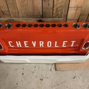 Op de afbeelding: Een rode en witte Chevrolet-thema bar met bekerhouders. De bar is ontworpen om de achterkant van een vintage Chevrolet-truck te lijken, met het woord "CHEVROLET" in witte letters. Het heeft meerdere bekerhouders en een witte bumper.