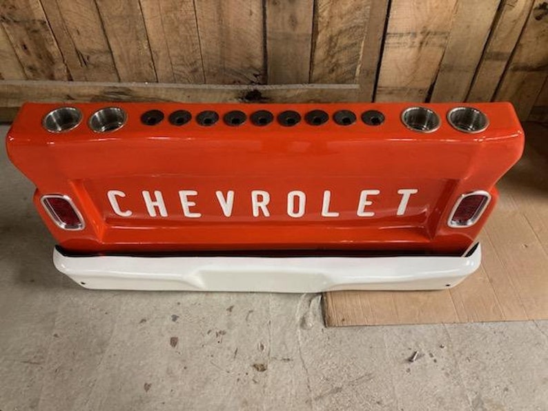 Op de afbeelding: Een rode en witte Chevrolet-thema bar met bekerhouders. De bar is ontworpen om de achterkant van een vintage Chevrolet-truck te lijken, met het woord "CHEVROLET" in witte letters. Het heeft meerdere bekerhouders en een witte bumper.