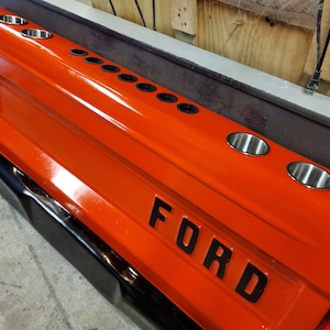 Op de afbeelding: Een oranje achterklep van een vrachtwagen met het woord "FORD" in zwarte letters. Het heeft meerdere roestvrijstalen bekerhouders. De achterklep heeft een zwarte bumper en een zilveren afwerking.