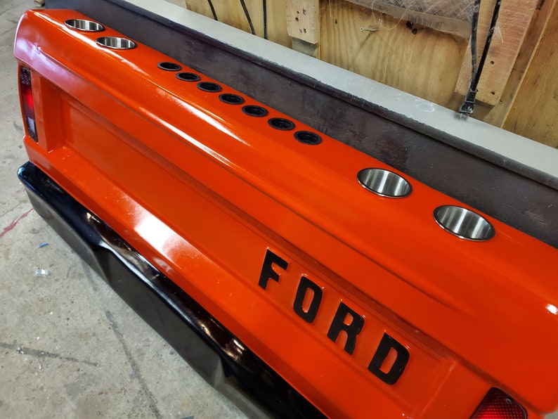 Op de afbeelding: Een oranje achterklep van een vrachtwagen met het woord "FORD" in zwarte letters. Het heeft meerdere roestvrijstalen bekerhouders. De achterklep heeft een zwarte bumper en een zilveren afwerking.