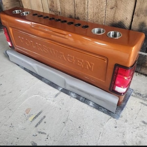 Op de afbeelding: Een op maat gemaakte, koperkleurige Volkswagen-truckachterklep met het woord "VOLKSWAGEN" in reli&euml;f in het midden. Het heeft ge&iuml;ntegreerde bekerhouders, achterlichten en een grijze bumper.