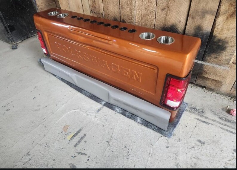 Op de afbeelding: Een op maat gemaakte, koperkleurige Volkswagen-truckachterklep met het woord "VOLKSWAGEN" in reli&euml;f in het midden. Het heeft ge&iuml;ntegreerde bekerhouders, achterlichten en een grijze bumper.