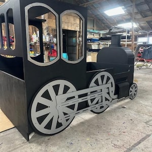 May include: A black wooden toy train engine with silver wheels. The train has a caboose with two windows. The train is sitting on a concrete floor.