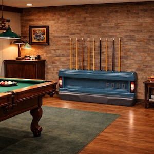 Op de afbeelding: Een biljartkamer met een pooltafel, een blauwe Ford-achterklep die als keurek wordt gebruikt en een bakstenen muur. De kamer wordt verlicht door groene hanglampen. De pooltafel heeft een groen vilt oppervlak.