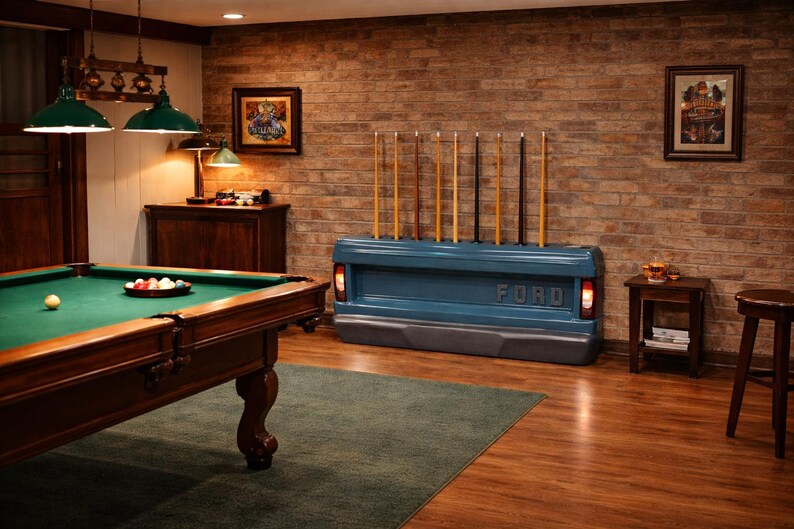 Op de afbeelding: Een biljartkamer met een pooltafel, een blauwe Ford-achterklep die als keurek wordt gebruikt en een bakstenen muur. De kamer wordt verlicht door groene hanglampen. De pooltafel heeft een groen vilt oppervlak.