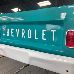 Op de afbeelding: Een turquoise Chevrolet-vrachtwagenachterklep met witte "CHEVROLET"-letters. De achterklep heeft een witte bumper en rode achterlichten. De afbeelding is een close-up van de achterkant van de vrachtwagen.