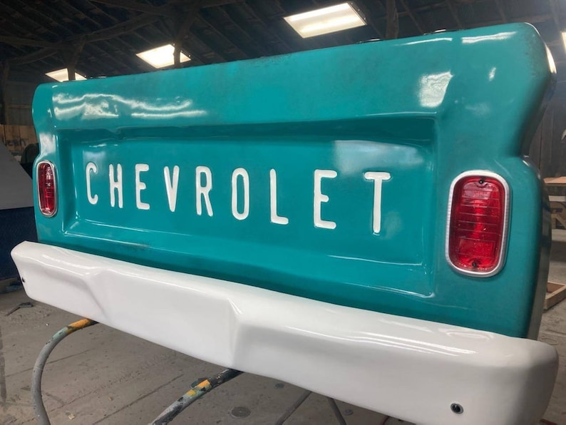 Op de afbeelding: Een turquoise Chevrolet-vrachtwagenachterklep met witte "CHEVROLET"-letters. De achterklep heeft een witte bumper en rode achterlichten. De afbeelding is een close-up van de achterkant van de vrachtwagen.