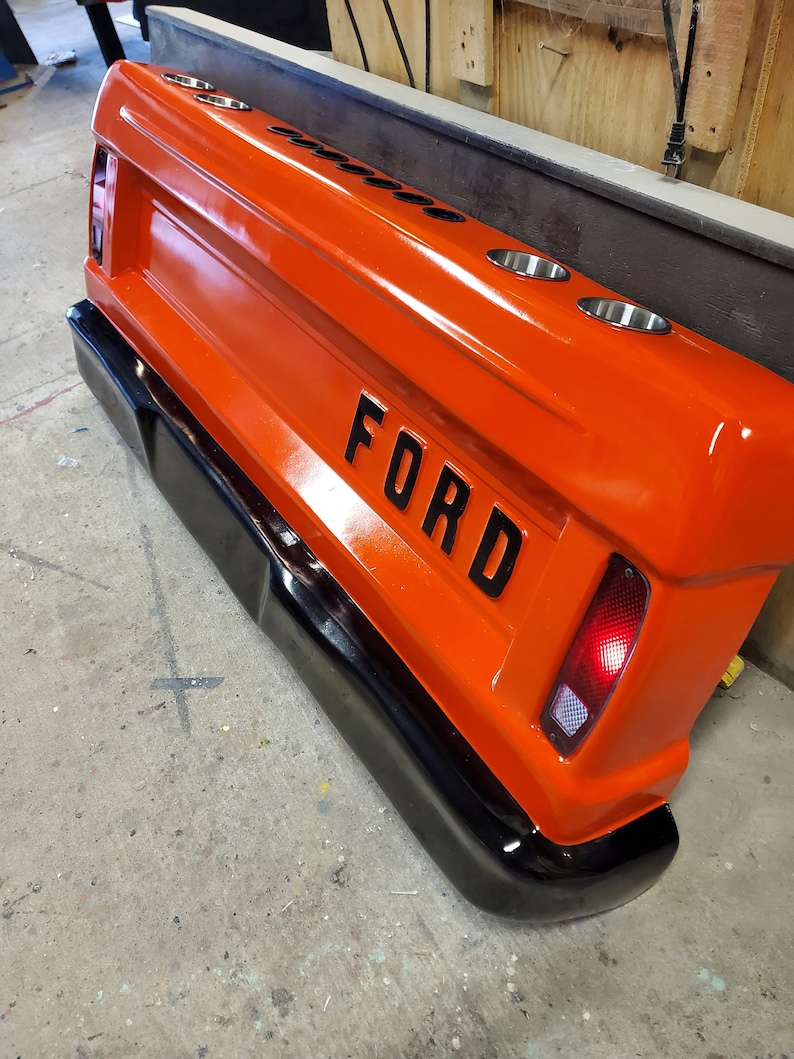 Op de afbeelding: Een oranje Ford-truckachterklep met het woord "FORD" in zwarte letters. De achterklep heeft vier bekerhouders en een zwarte bumper. Het achterlicht is aan de rechterkant zichtbaar.