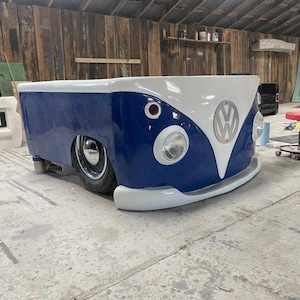 May include: A blue and white Volkswagen bus bed frame. The bed frame is made to look like the front of a classic Volkswagen bus. The bed frame has a white front bumper and a white roof. The Volkswagen logo is visible on the front of the bed frame.