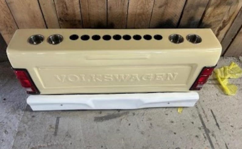 Op de afbeelding: Beige op maat gemaakte achterklep met het woord "VOLKSWAGEN" in reli&euml;f. Het heeft meerdere bekerhouders en een witte bumper eronder. Rode achterlichten zijn zichtbaar.