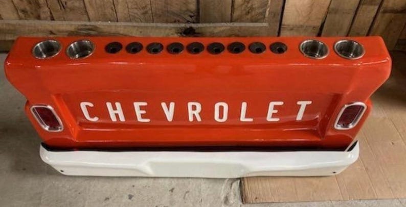 Op de afbeelding: Een oranje Chevrolet achterklepbar met het woord "CHEVROLET" in het wit. Het heeft verschillende bekerhouders en een witte bumper. De bar is ontworpen om op de achterkant van een klassieke pick-up truck te lijken.