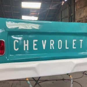 Op de afbeelding: Een turquoise Chevrolet-achterklep met het woord "CHEVROLET" in witte letters. De achterklep heeft een witte bumper en rode achterlichten. Klassiek ontwerp van een vintage pick-up.