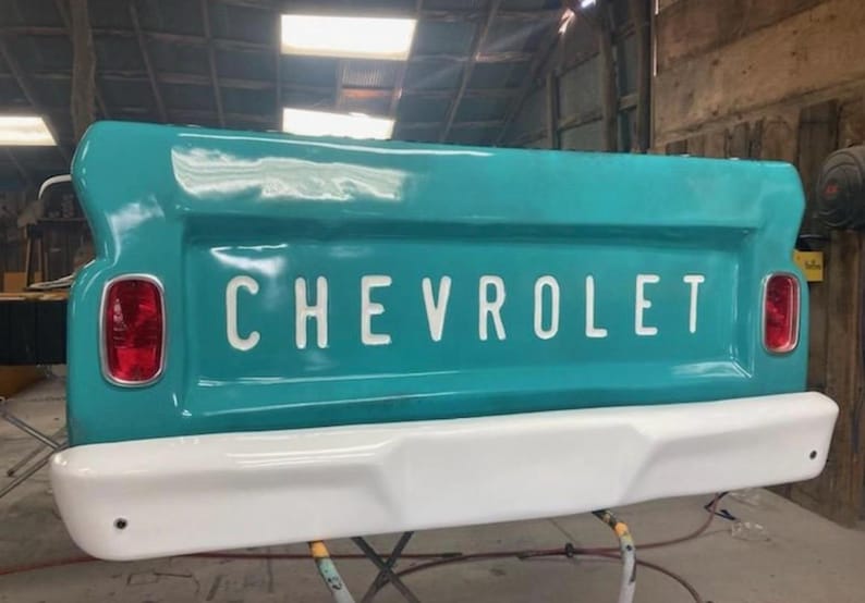 Op de afbeelding: Een turquoise Chevrolet-achterklep met het woord "CHEVROLET" in witte letters. De achterklep heeft een witte bumper en rode achterlichten. Klassiek ontwerp van een vintage pick-up.