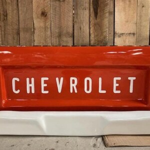 Op de afbeelding: Een felrode Chevrolet-truckachterklep met de merknaam in witte blokletters. De achterklep heeft een witte bumper en twee rode achterlichten. De achtergrond is een houten muur.