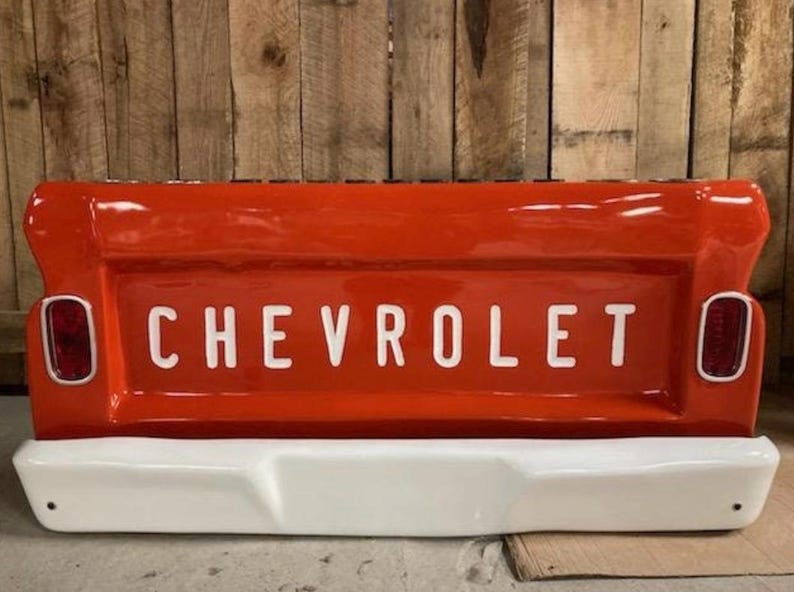 Op de afbeelding: Een felrode Chevrolet-truckachterklep met de merknaam in witte blokletters. De achterklep heeft een witte bumper en twee rode achterlichten. De achtergrond is een houten muur.