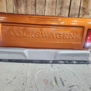 Op de afbeelding: Oranje Volkswagen-vrachtwagenachterklep met het woord "VOLKSWAGEN" in reli&euml;f. De achterklep heeft een grijze bumper en rode achterlichten. De achtergrond is een houten muur en een betonnen vloer.