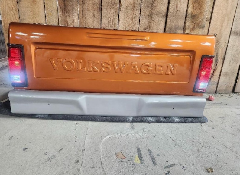 Op de afbeelding: Oranje Volkswagen-vrachtwagenachterklep met het woord "VOLKSWAGEN" in reli&euml;f. De achterklep heeft een grijze bumper en rode achterlichten. De achtergrond is een houten muur en een betonnen vloer.