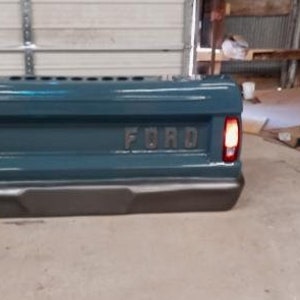 Op de afbeelding: Tealgekleurde laadbak met het woord "FORD" in reli&euml;f in het midden. De laadbak heeft een donkergrijze bumper en rode achterlichten. De foto is binnenshuis gemaakt.