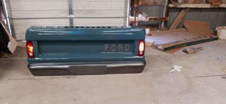 Op de afbeelding: Tealgekleurde laadbak met het woord "FORD" in reli&euml;f in het midden. De laadbak heeft een donkergrijze bumper en rode achterlichten. De foto is binnenshuis gemaakt.