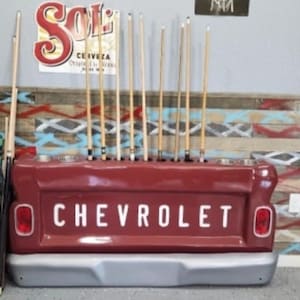 Op de afbeelding: Een rode Chevrolet-vrachtwagenbak die is omgebouwd tot een biljartkeu-rek. Het rek toont het woord "CHEVROLET" in witte letters. De achtergrond toont een kleurrijke, patroonmuur en een "SOL"-bord, met keuen.