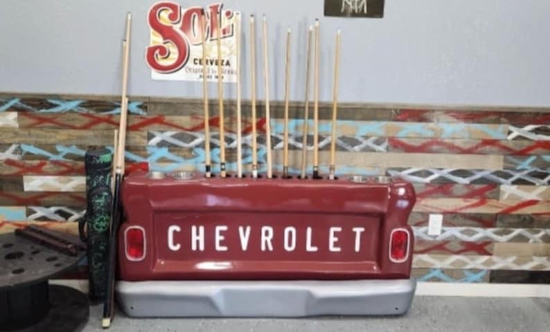 Op de afbeelding: Een rode Chevrolet-vrachtwagenbak die is omgebouwd tot een biljartkeu-rek. Het rek toont het woord "CHEVROLET" in witte letters. De achtergrond toont een kleurrijke, patroonmuur en een "SOL"-bord, met keuen.