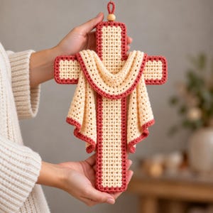 May include: A crochet cross wall hanging featuring a cream-colored base with a dark red border. A cream-colored fabric drape with a dark red edge is layered over the center of the cross. A small wooden bead sits at the top hanging loop.