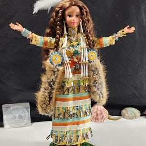 Op de afbeelding: Pop gekleed in inheemse Amerikaanse kledij, met een gedetailleerde gele, oranje en turquoise jurk met franjes. De pop heeft lang bruin haar, een gevederde hoofdtooi en staat op groen mos. Accessoires zijn een bontstola en kralenjuwelen.
