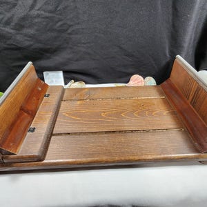 May include: A wooden serving tray with a dark brown finish. The tray has a rectangular shape with raised sides and a slatted base. The sides are angled and have a lighter wood trim. The tray is designed to be foldable.