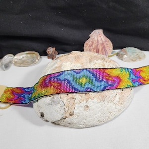 May include: A colorful beaded bracelet with a vibrant rainbow pattern. The bracelet features a mix of pink, blue, yellow, and green beads, with tan leather ends and a tan leather cord. The bracelet is displayed on a white surface.