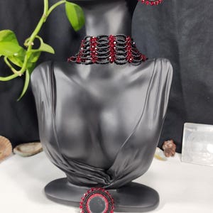 Può includere: Un girocollo nero con perline e dettagli in cristallo rosso. Sono anche raffigurati orecchini e un anello abbinati in cristallo rosso e perline nere.