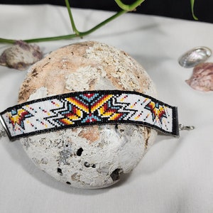 May include: A white beaded bracelet with a geometric pattern in red, yellow, blue, and black. The bracelet has silver metal clasps.