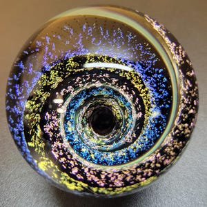 May include: A spherical glass paperweight featuring a deep black base with a swirling vortex pattern. The surface is embedded with iridescent flecks of blue, gold, and purple that create a shimmering, galaxy-like effect within the clear glass.