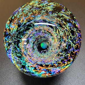 May include: A circular glass paperweight featuring a deep, swirling vortex design. The clear glass is embedded with iridescent, multi-colored metallic flakes in shades of blue, green, gold, and purple, creating a galaxy-like effect around a central point.