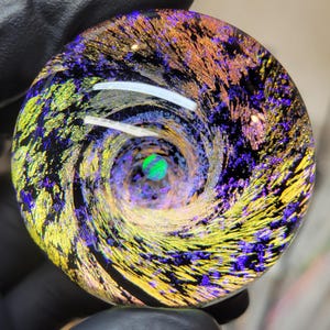 May include: A circular glass pendant featuring a swirling vortex pattern of iridescent gold, purple, and black colors. At the center of the spiral sits a small, round, bright green opal-like inlay that creates a focal point within the glass.