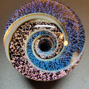 May include: A round, glass paperweight with a spiral design. The center is a deep black, surrounded by a swirling pattern of blue, gold, and purple. The outer edge has a clear, iridescent band.