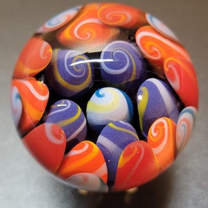 May include: A glass paperweight with a swirling pattern of orange, white, and purple. The paperweight is filled with smaller glass marbles of various colors.