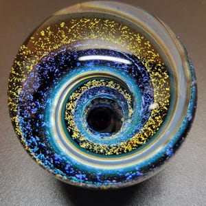 May include: A circular glass object featuring a deep spiral pattern that transitions from a dark center to outer rings. The surface is decorated with iridescent blue and gold flecks that create a shimmering, galaxy-like effect across the glass.