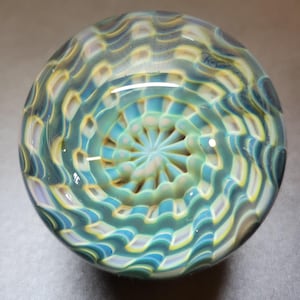 May include: A round, glass art piece with a swirling, patterned design. The colors include shades of teal, yellow, and white, creating a mesmerizing effect. The center features a starburst pattern. The glass is smooth and reflective.