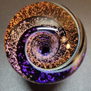 Pode incluir: Um globo de vidro transparente com um design espiral interno. O globo apresenta um padrão semelhante a uma galáxia com manchas laranja e roxas, e um vórtice central. A borda externa tem um redemoinho azul claro e branco.