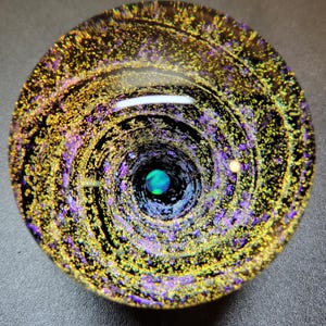 May include: A round glass paperweight with a swirling galaxy design. The paperweight features a dark background with gold, purple, and blue colors. The center has a small blue orb.