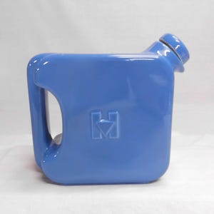 May include: A glossy, cornflower blue ceramic pitcher shaped like a square fuel canister. It features a side handle, a slanted spout with a matching blue cap, and a raised, embossed letter H with a downward-pointing triangle centered on the side.