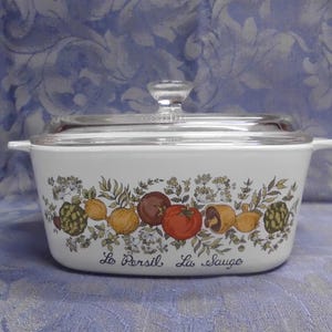May include: White rectangular casserole dish with a clear glass lid. The dish is decorated with a colorful vegetable and herb design, including tomatoes, mushrooms, and artichokes. The words "Le Persil La Sauge" are written on the front.