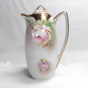 May include: A white porcelain pitcher with a lid, featuring a hand-painted design of pink roses and green leaves. The handle and rim are trimmed with gold accents. A decorative piece for serving beverages.