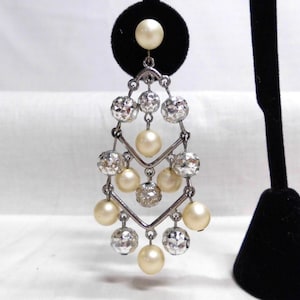 May include: A chandelier earring featuring a silver-toned metal frame adorned with alternating cream-colored faux pearls and clear, faceted, spherical beads arranged in a geometric, multi-tiered drop design with a single pearl stud top.