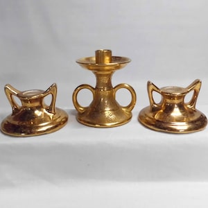 May include: A set of three gold-colored candle holders. The set includes two small, low candle holders with curved handles and a taller, central candle holder with a round base and two loop handles. The candle holders have a textured surface.