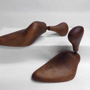 May include: Two antique wooden shoe stretchers, in a warm brown hue, are displayed against a white backdrop. The stretchers are designed to maintain the shape of shoes. One is partially disassembled, showcasing its adjustable mechanism.