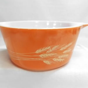 May include: An orange Pyrex bowl with a white interior and two handles. The exterior features a wheat pattern in a lighter orange color. This vintage kitchenware is ideal for baking and serving.