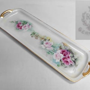 May include: A rectangular porcelain serving tray with rounded corners and gold-trimmed handles. The white surface features a hand-painted floral motif of pink and white roses with green leaves. A maker's mark reads R.S. Germany with a star and wreath.
